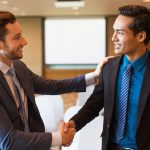 Closeup of two smiling middle-aged business men shaking hands and greeting each other in conference room with whiteboard in background